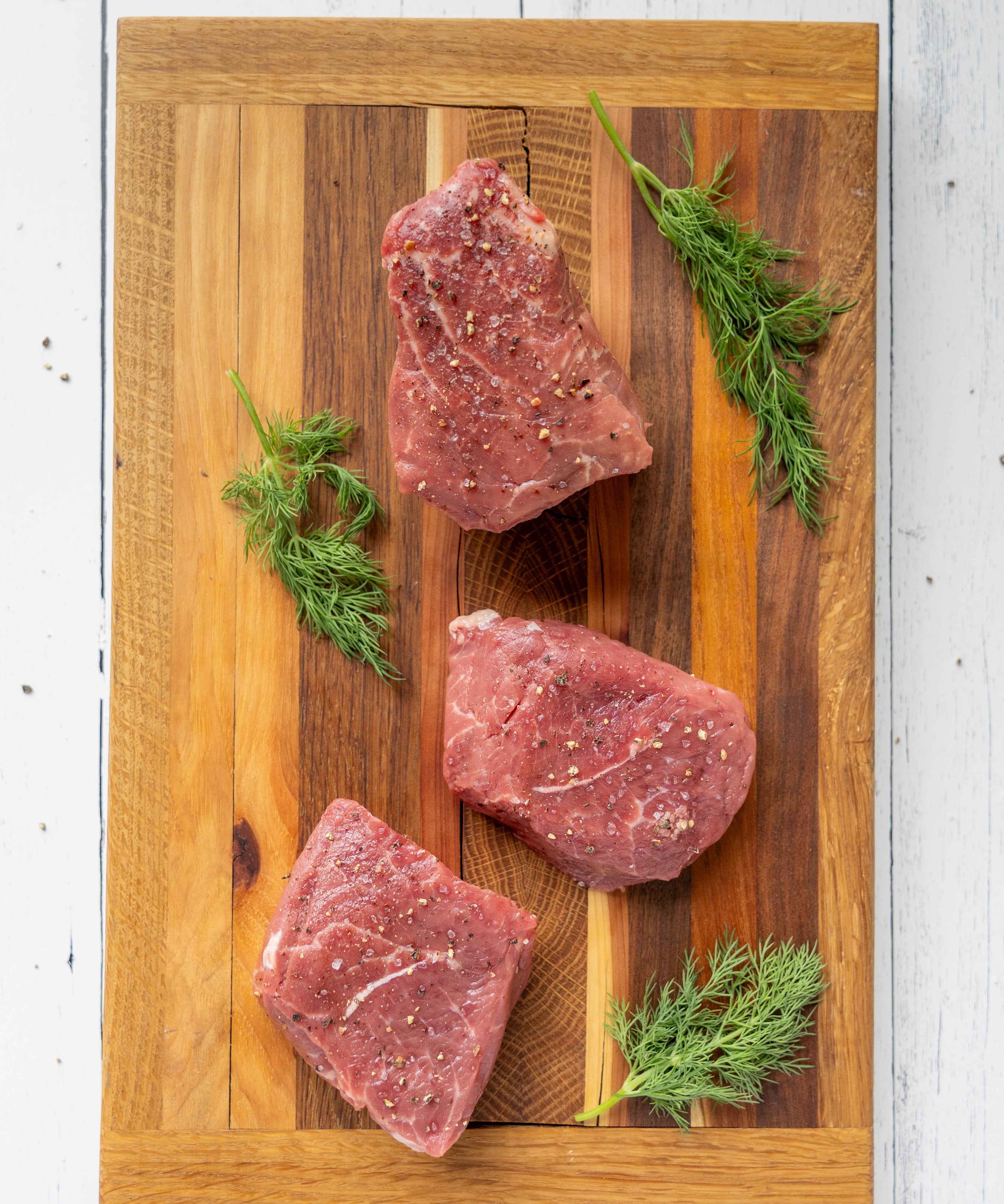 Three pieces of raw meat on a wooden cutting board with fresh herbs.
